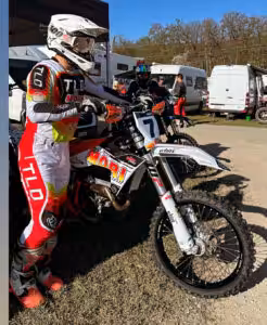 Motocross-Fahrer in bunter Ausrüstung und Helm steht mit Motorrad in einem Rennlager.