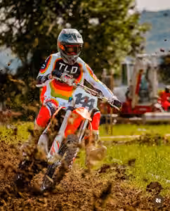 Motocrossfahrer auf rotem Motorrad in Aktion, wirbelt Schlamm auf, umgeben von Natur und hindurch stürmendem Gelände.