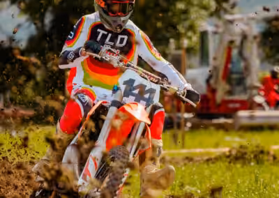 Motocrossfahrer auf rotem Motorrad in Aktion, wirbelt Schlamm auf, umgeben von Natur und hindurch stürmendem Gelände.