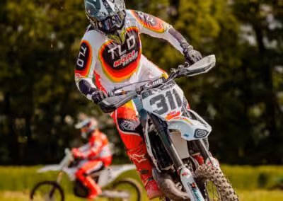 Motocross-Fahrer springt über Hügel auf Rennstrecke im Wald, actionreiches Offroad-Abenteuer bei sonnigem Wetter.