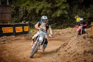 Motocrossfahrer bei einem Rennen auf schlammiger Piste, umgeben von Bäumen und Pirelli-Werbung im Hintergrund.